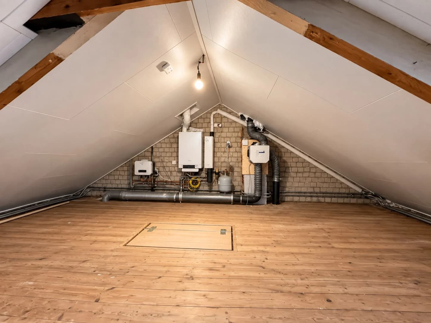 Attic with white sloping finishes, a timber floor hatch and existing services on the gable wall