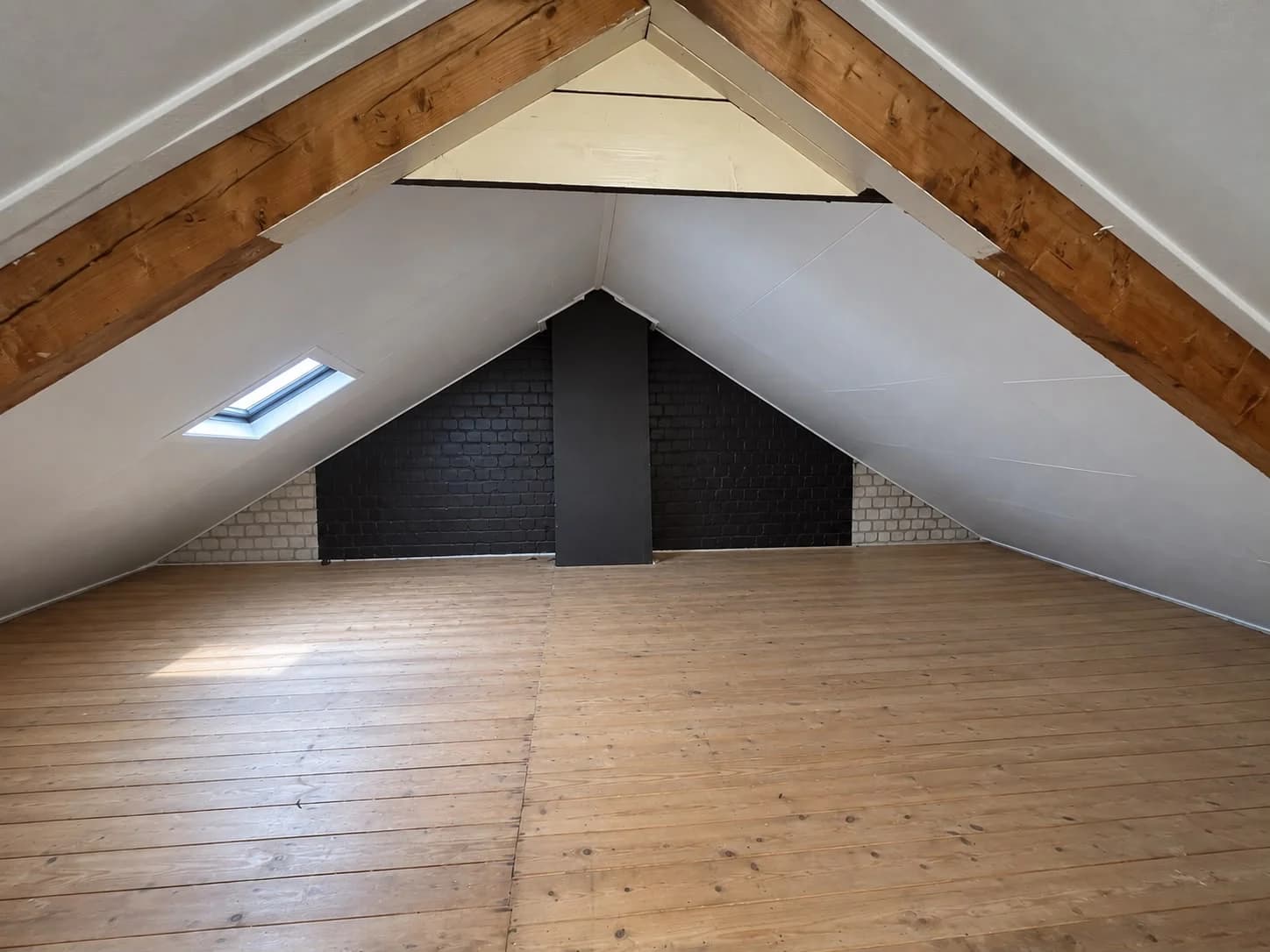 Attic with white sloping panels, timber beams, a dark gable wall and a roof window on the left