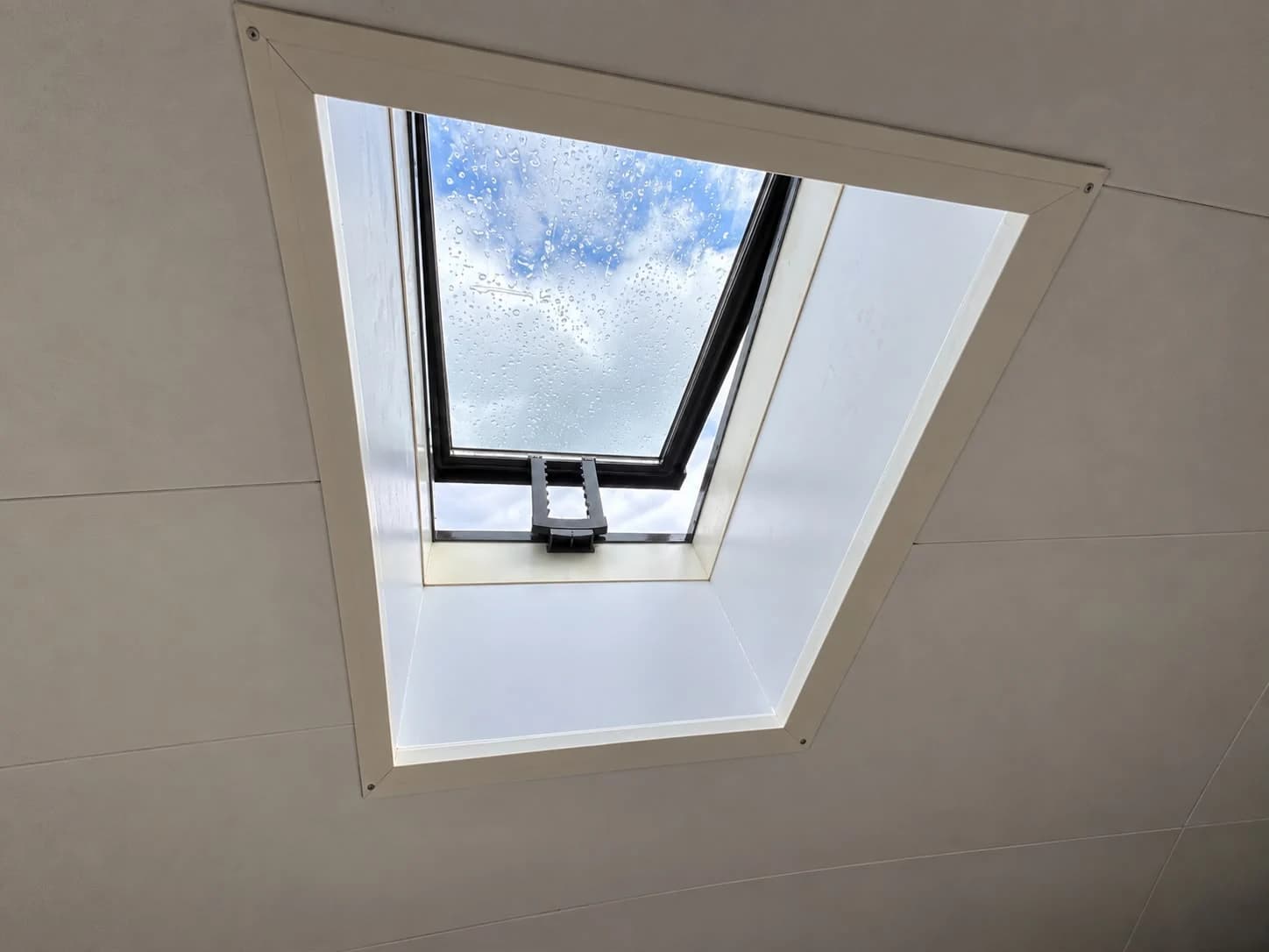 Roof window with a dark frame edge, white interior lining and raindrops on the glass